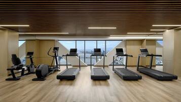 Een fitnessruimte met loopbanden, elliptische trainers en een roeimachine. Er ligt een houten vloer en er is uitzicht op een stadsgezicht door grote ramen.