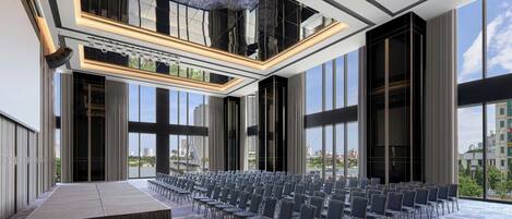 Un salón de conferencias amplio con filas de sillas, un escenario y grandes ventanales que ofrecen vistas a la ciudad.
