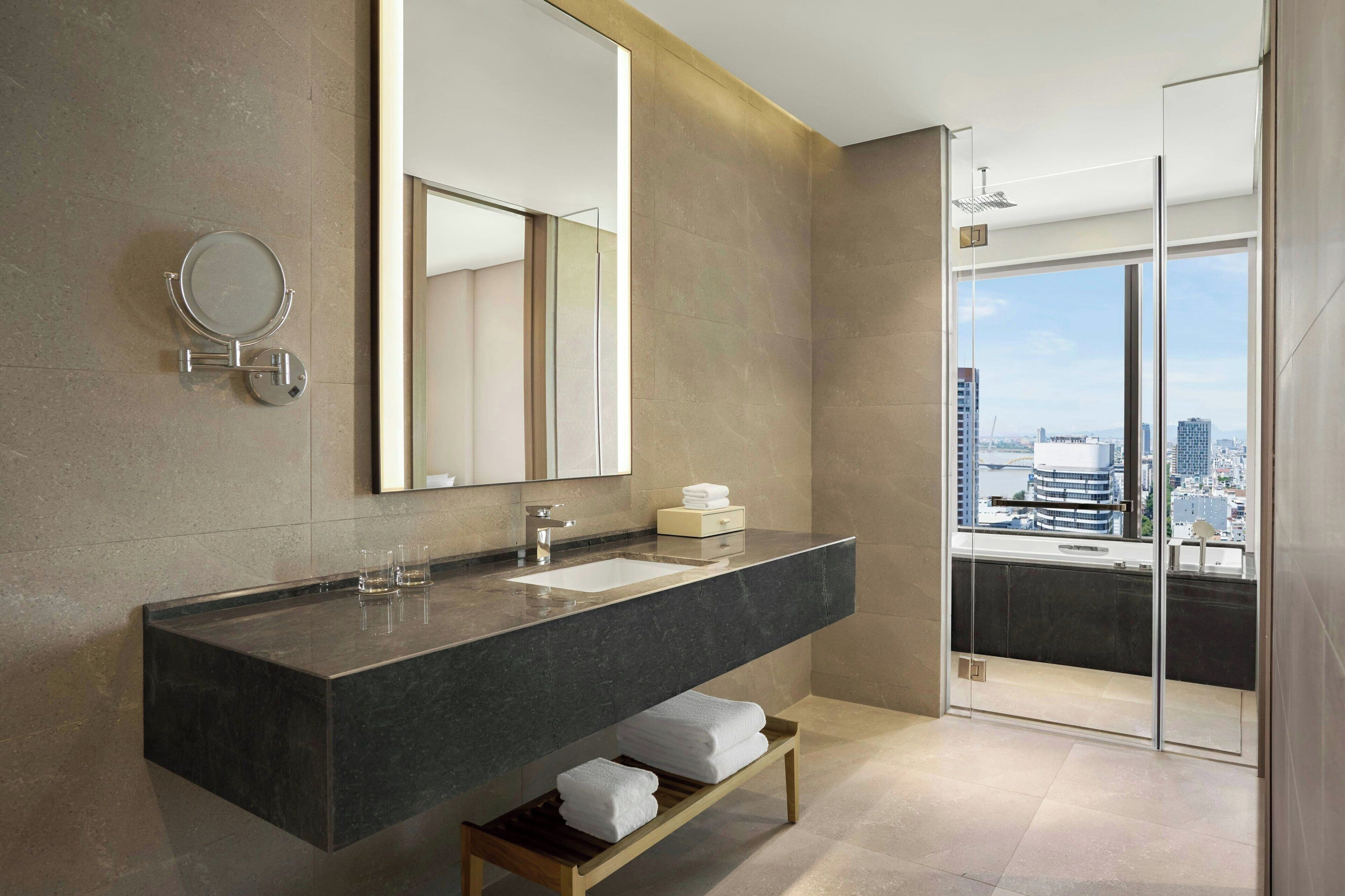 Une salle de bain moderne avec un grand miroir, un plan de toilette en marbre foncé, une douche à parois de verre et une vue sur la ville.