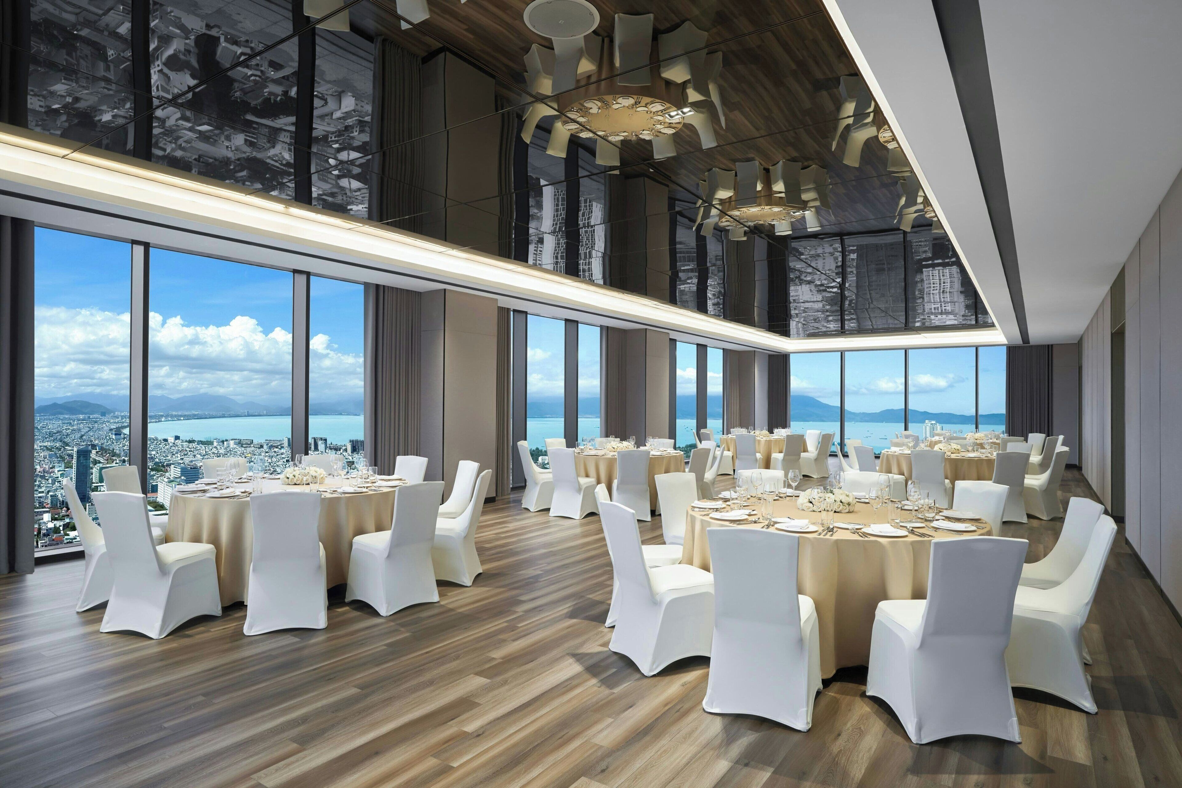 A spacious conference room with round tables, white chairs, and large windows offering a city and ocean view.