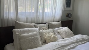 A neatly made bed with white bedding and fluffy pillows, a window with sheer curtains, and a wall-mounted fan. - Darling Bungalow in the heart of quaint Inglewood, On. 15 minutes from Osprey! (Inglewood)