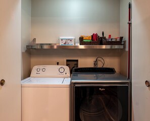 Un espacio reducido para lavandería con lavadora Amana y secadora Whirlpool, un estante con productos de limpieza y una cesta con artículos de limpieza.