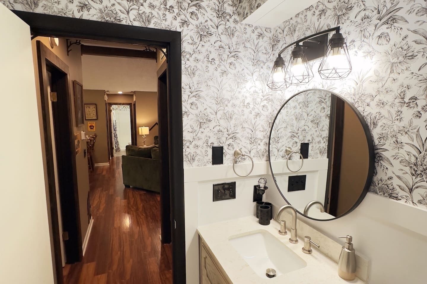 A bathroom with a large mirror, a sink, and a shower. There is a floral wallpaper and a wooden floor.