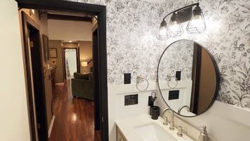 A bathroom with a large mirror, a sink, and a shower. There is a floral wallpaper and a wooden floor.