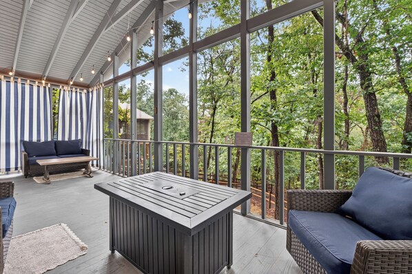 A covered outdoor area with a table and seating, large windows, and a view of trees.