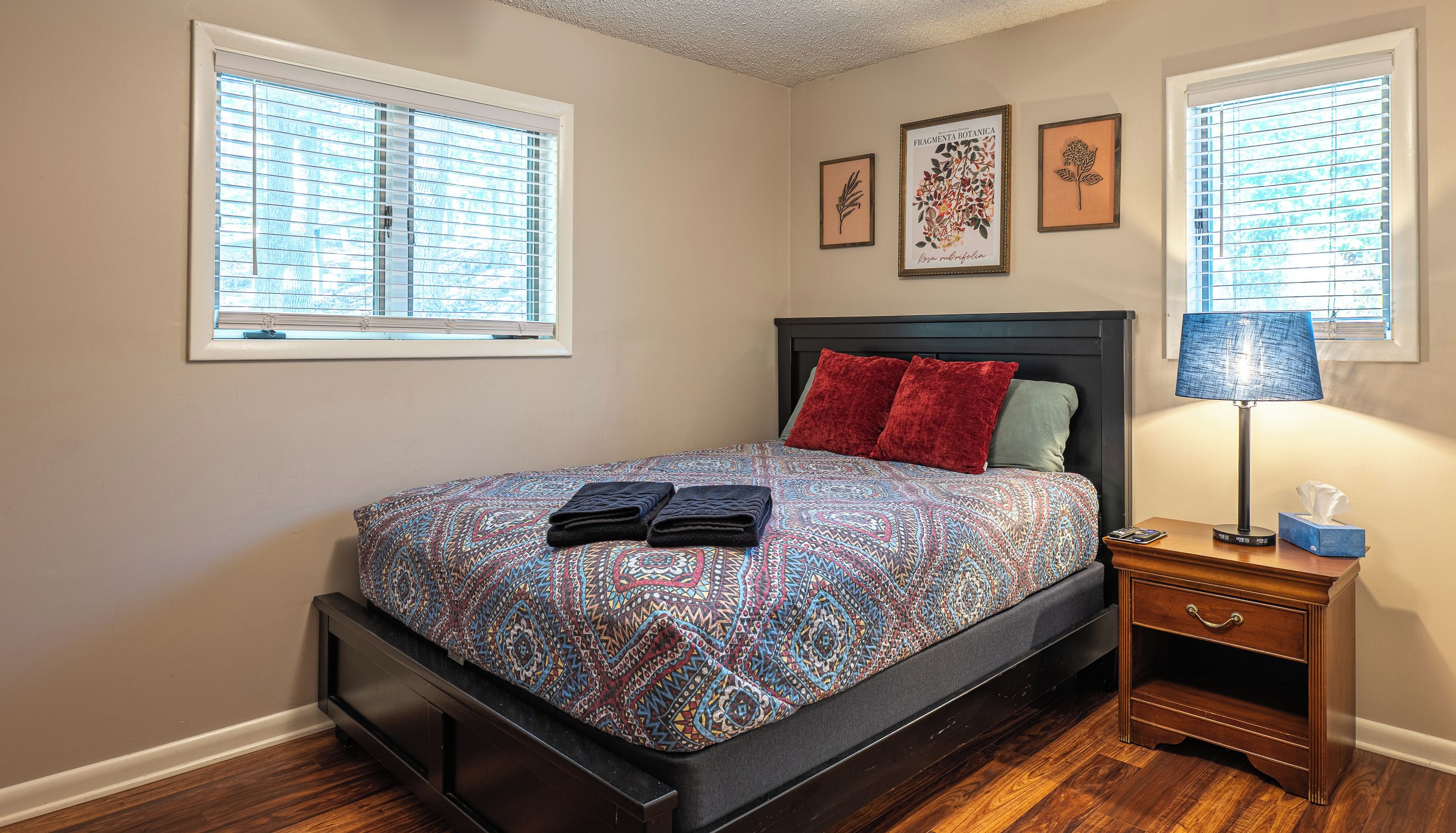 A bedroom with a bed, a nightstand, a lamp, and two windows with blinds.