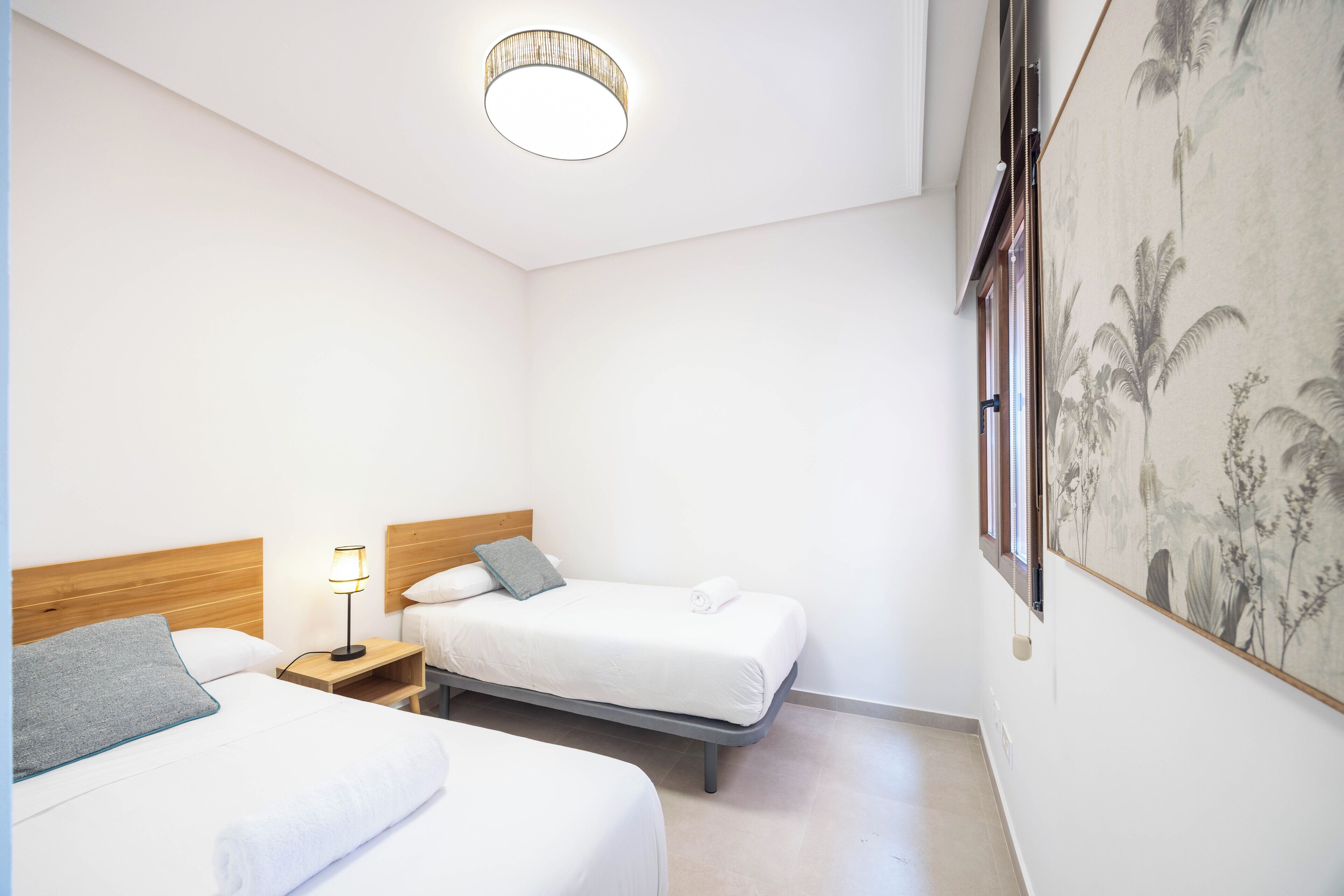 A hotel room with two beds, a nightstand with a lamp, and a large window with a view of palm trees.