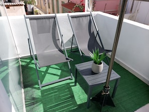 Un balcón con piso de césped artificial verde, dos sillas de malla grises, una mesita con una planta en maceta y un poste de metal.