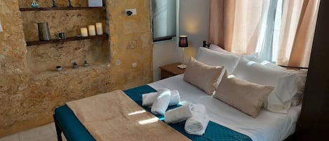 Una habitación de hotel con pared de piedra, cama con ropa de cama azul y blanca, mesita de noche con lámpara y repisas con objetos decorativos.