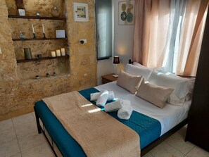 A hotel room with a stone wall, a bed with blue and white bedding, a nightstand with a lamp, and a shelf with decorative items.