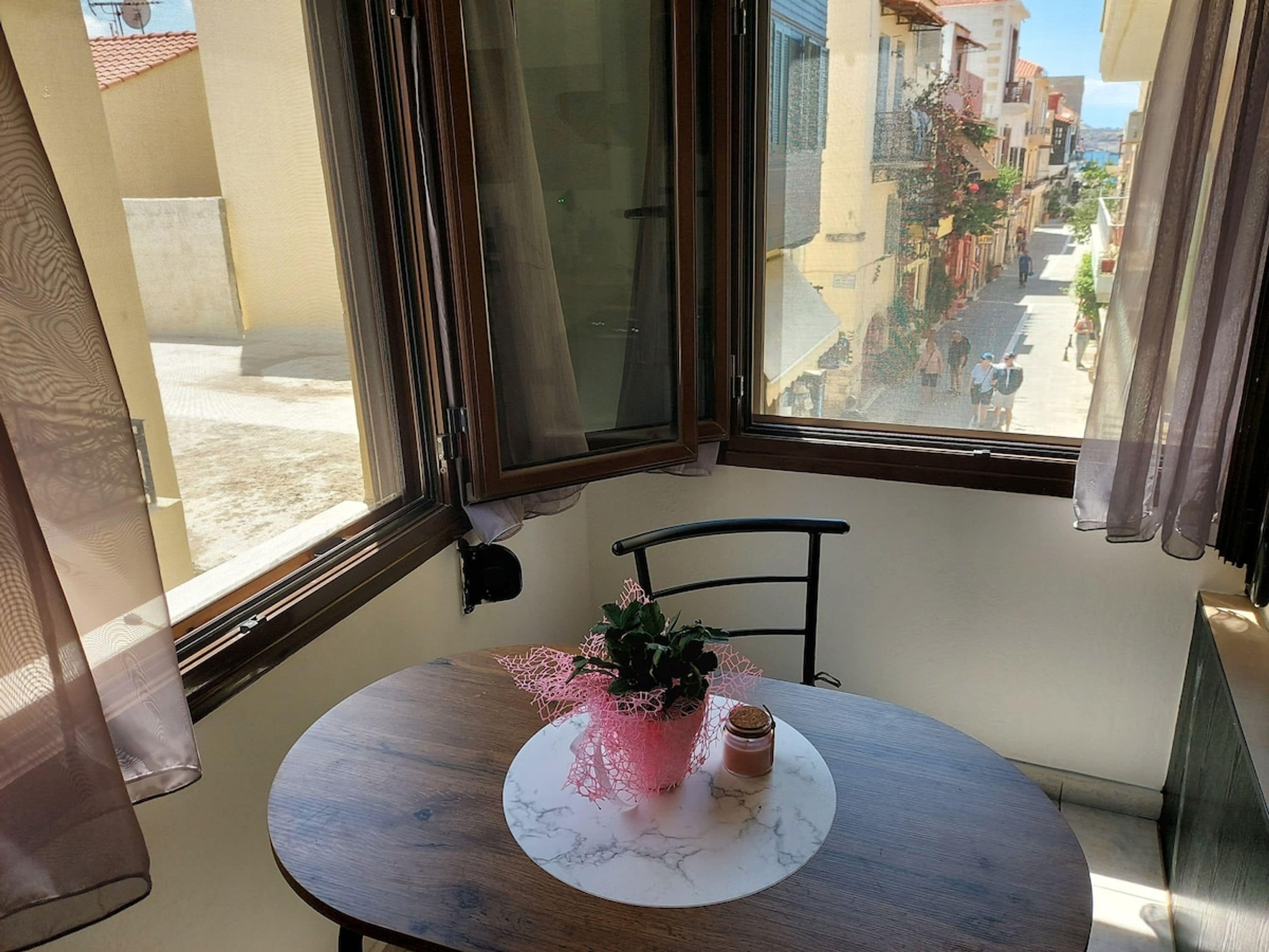A room with a round table, a chair, a window with curtains, and a view of a street with buildings.