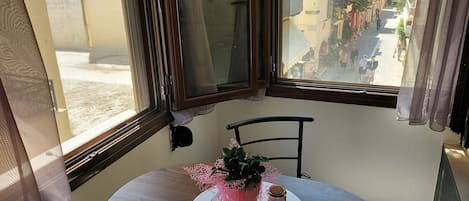 A room with a round table, a chair, a window with curtains, and a view of a street with buildings.