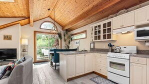 A modern kitchen with wooden beams, white cabinets, a built-in oven, and a large window overlooking greenery. - The Crow's Nest Suite - Steps from S. Chesterman (Tofino)