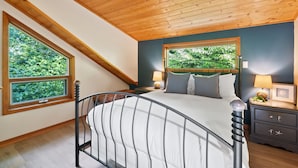 A bedroom with a sloped ceiling, a large window, a bed with a headboard, bedside tables, and a nightstand with a lamp. - The Crow's Nest Suite - Steps from S. Chesterman (Tofino)