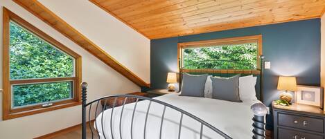 Une chambre avec un plafond en pente, une grande fenêtre, un lit avec une tête de lit, des tables de chevet et une table de nuit avec une lampe.