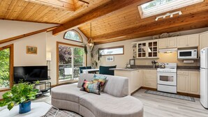 A modern kitchen with wooden cabinets, a microwave, and a stove. There is a large window providing natural light and a view of greenery outside. - The Crow's Nest Suite - Steps from S. Chesterman (Tofino)