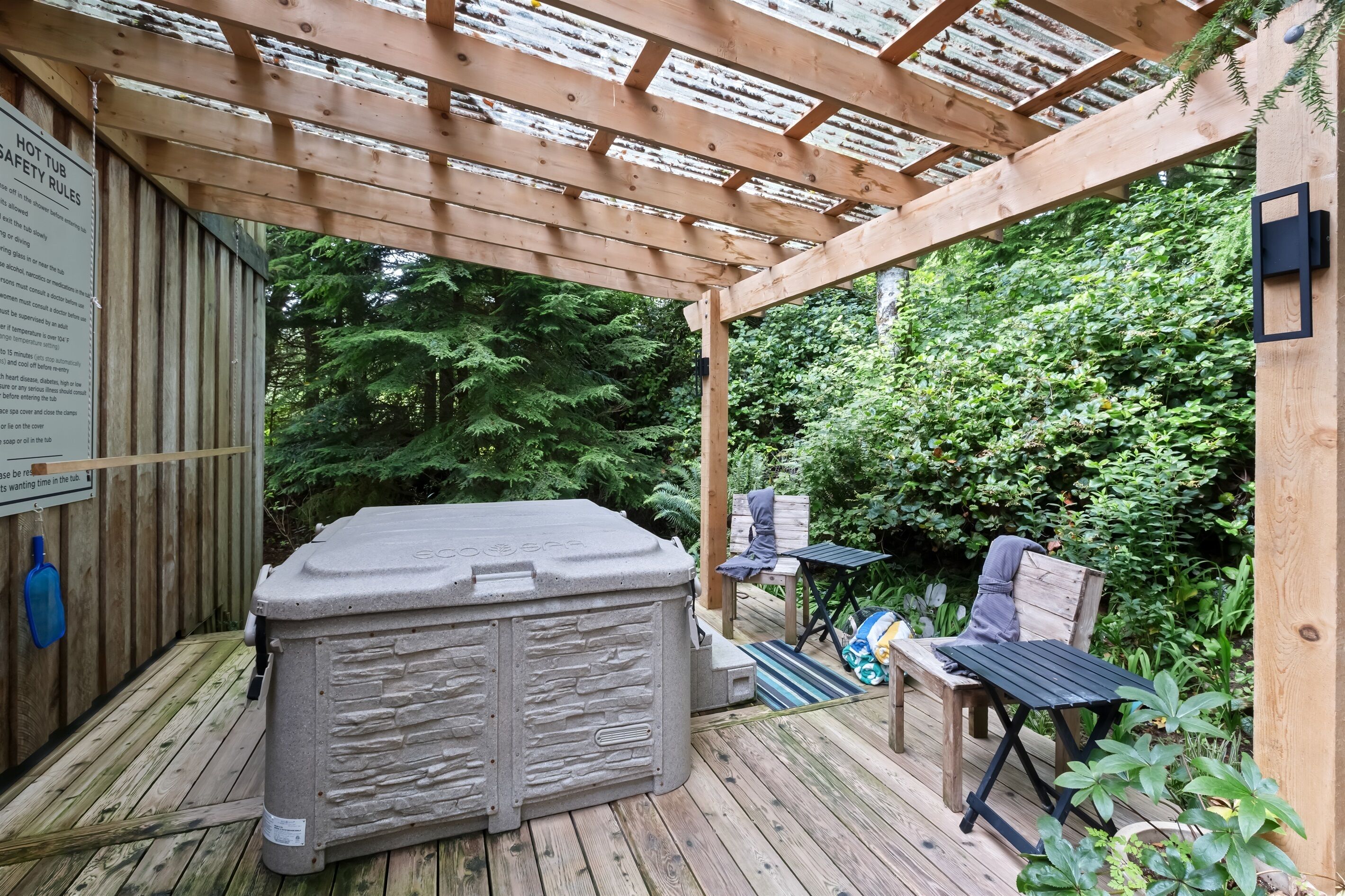 A hot tub under a wooden pergola with a safety rule sign, surrounded by lush greenery.