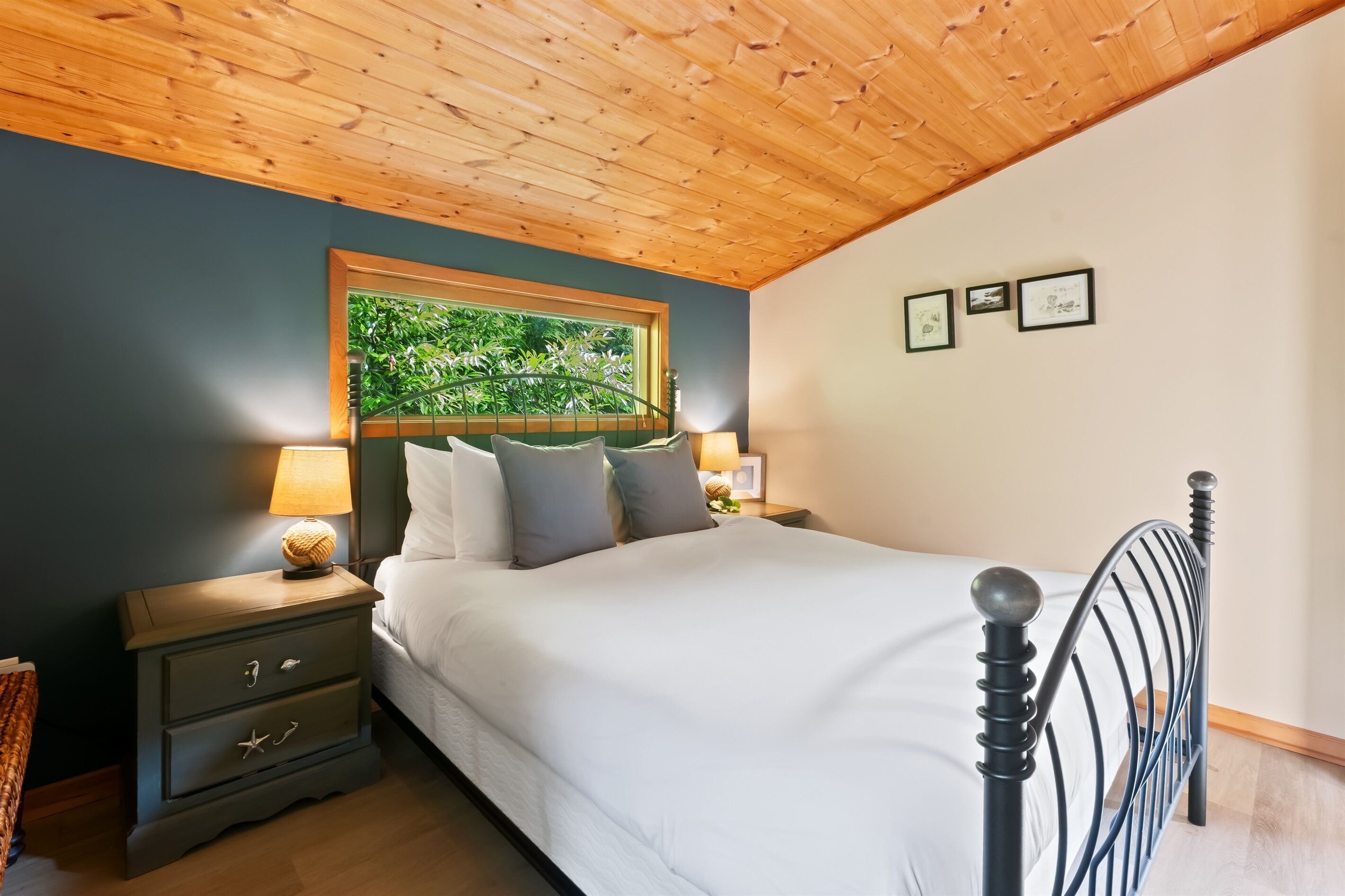 A bedroom with a wooden ceiling, a bed with white bedding, a nightstand with a lamp, and a window with a view of greenery.