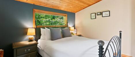 A bedroom with a wooden ceiling, a bed with white bedding, a nightstand with a lamp, and a window with a view of greenery.