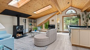 A modern loft-style living room with a fireplace, a curved sofa, a dining area, and a kitchen with a large window. - The Crow's Nest Suite - Steps from S. Chesterman (Tofino)