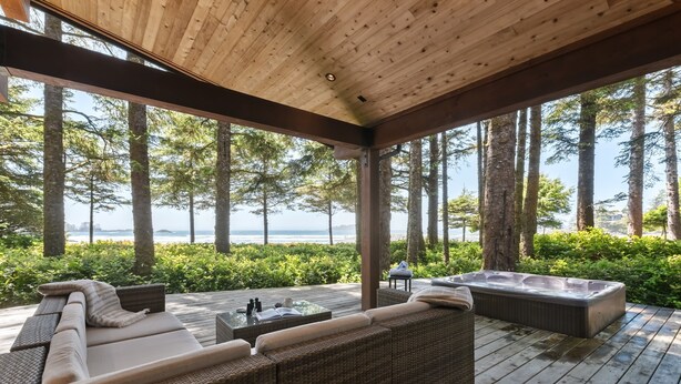 A covered outdoor seating area with wicker furniture and a hot tub, overlooking a forested beach.