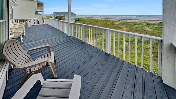 A wooden deck with chairs, a view of the beach, and a house in the background. - Whispering Sands Beachfront | Community Pool (Galveston)