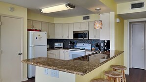 A kitchen with white cabinets, a granite countertop, a microwave, a stove, and a sink. There are three wooden stools at the island. - Whispering Sands Beachfront | Community Pool (Galveston)