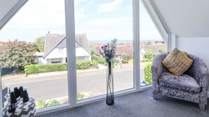 Una habitación con un ventanal que ofrece vistas a una calle y casas. Hay un sofá gris con un cojín amarillo y un jarrón con flores sobre una mesa.