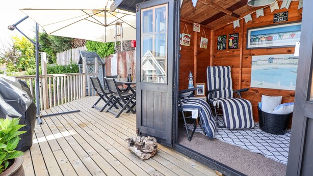 Una terraza de madera con mobiliario exterior, una parrilla y un área cubierta con sillas a rayas y una mesa.