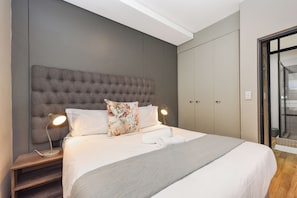 A modern bedroom with a tufted headboard, two bedside lamps, a neatly made bed with white linens, and a large wardrobe.