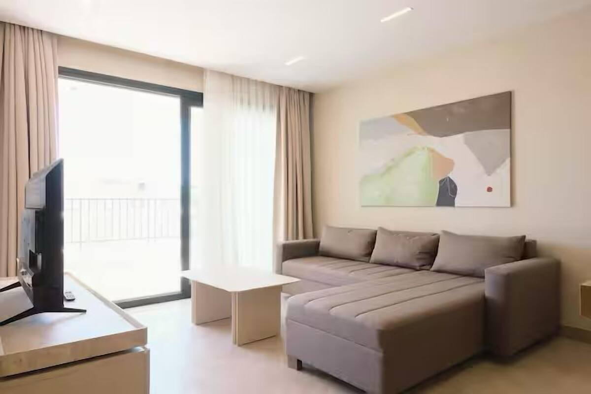 A modern hotel room with a large bed, a flat-screen TV, a small table, and a large window with curtains.