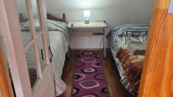 A small, narrow room with two beds, a desk with a lamp, and a patterned rug.
