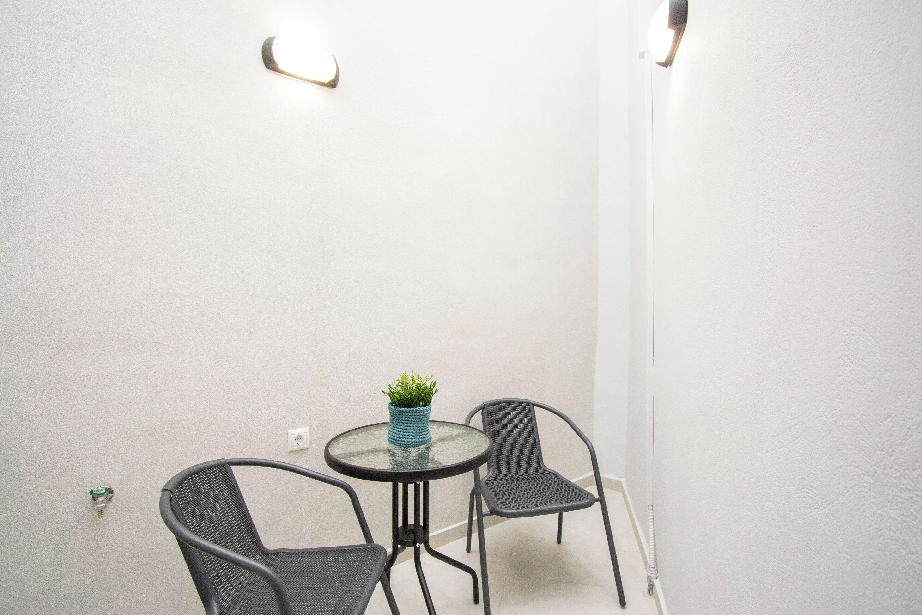 Een kleine, ronde glazen tafel met twee zwarte metalen stoelen, een kleine blauwe vaas met groene planten en twee wandlampen.