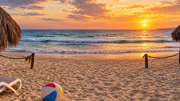 A beach with a rope barrier, a beach ball, and a thatched umbrella.