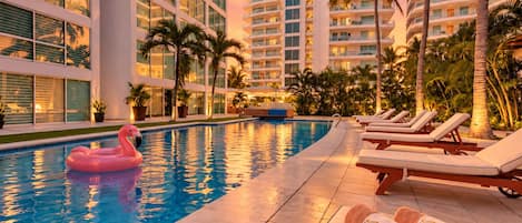 Una piscina con sillas de descanso y un flotador con forma de flamenco, rodeada de palmeras y edificios modernos.