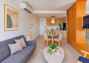 Una sala de estar moderna con un sofá gris, una mesa de centro con una planta, y un comedor con sillas de madera y una mesa preparada para cuatro personas.