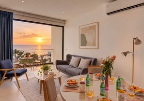 Una sala de estar moderna con un sofá gris, una mesa redonda de centro y una mesa de comedor dispuesta con platos, vasos y botellas. Un ventanal ofrece vistas de la puesta de sol sobre el mar.