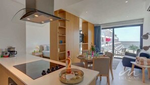 A modern kitchen with a central island, a dining area with a table and chairs, and a view of the sea through large windows.