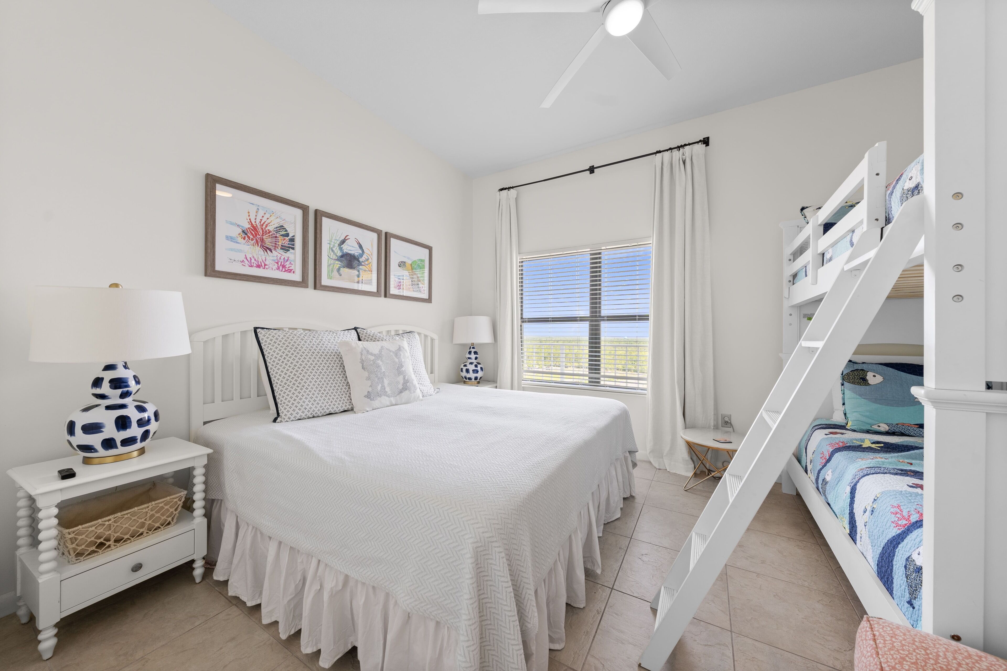 A bedroom with a bunk bed, a white bedspread, a nightstand with a lamp, and framed pictures on the wall.