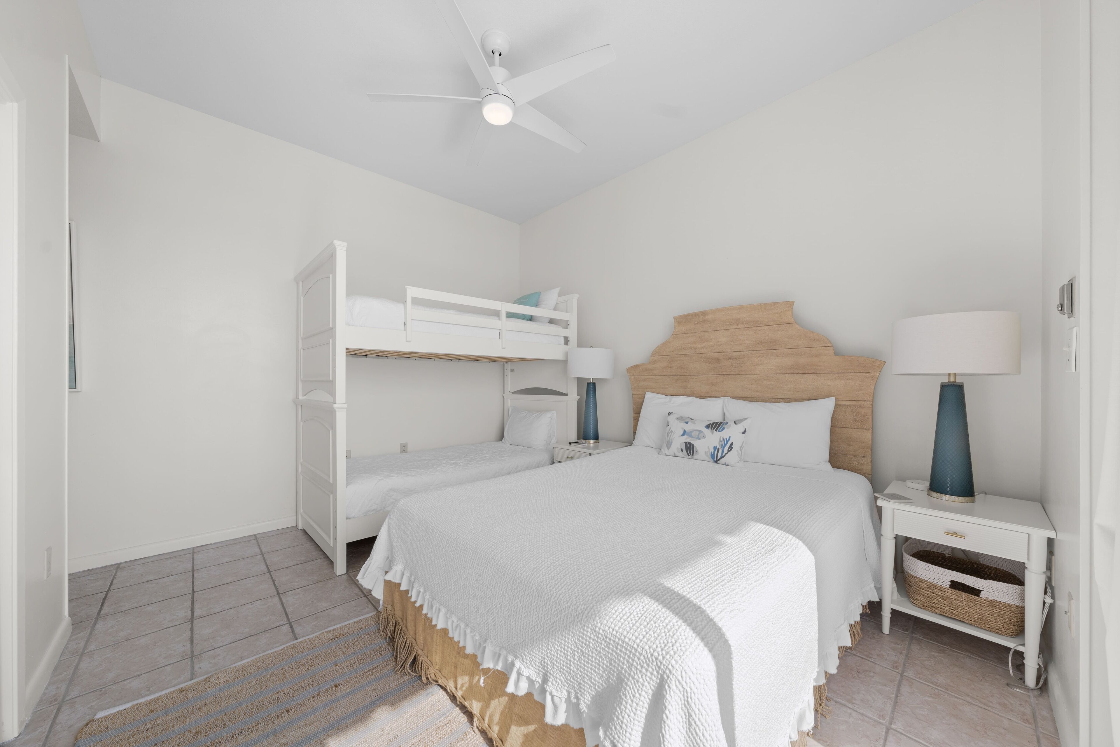 A bedroom with a bunk bed, a single bed, a wooden headboard, two bedside tables with lamps, and a ceiling fan.