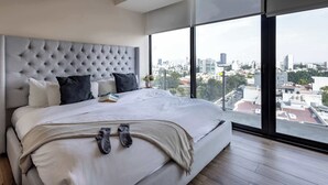 A modern hotel room with a large bed, a grey tufted headboard, and a city view through large windows.