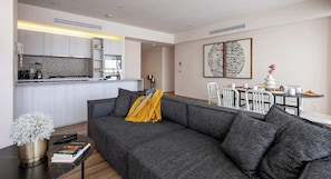 A modern living area with a grey sofa, a small table with books, a decorative plant, and a kitchen in the background.