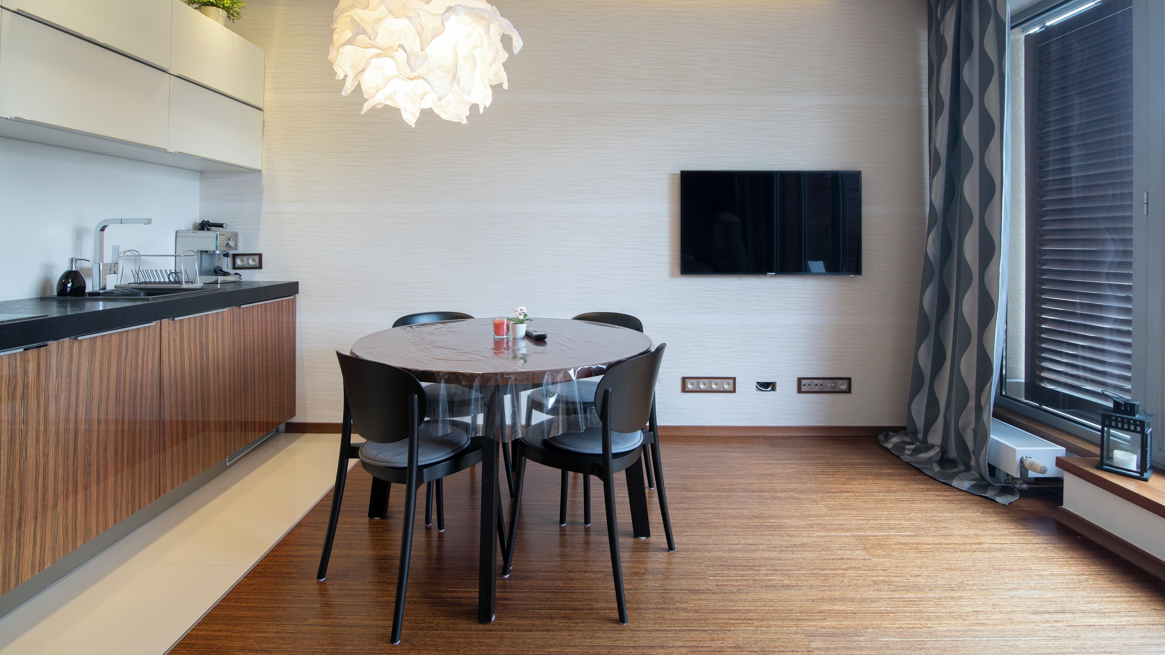 Un comedor moderno con una mesa redonda, cuatro sillas, una lámpara colgante, un televisor de pantalla plana en la pared y una encimera de cocina con electrodomésticos.
