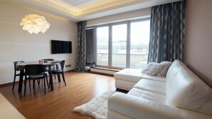 Un salon moderne avec un canapé blanc, une table à manger et des chaises. On y trouve une grande fenêtre avec des rideaux, un téléviseur à écran plat fixé au mur et un tapis moelleux sur le sol.