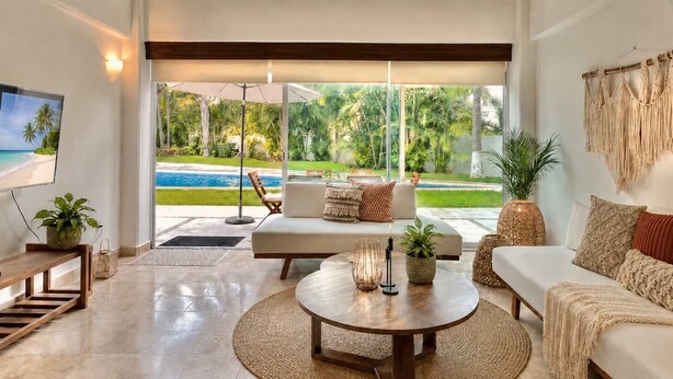 Ein großzügiges Wohnzimmer mit einem großen Fenster mit Blick auf einen Pool und Palmen, einem Deckenventilator und einem Fernseher an der Wand.