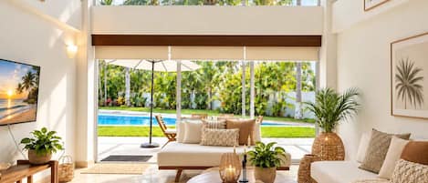 A spacious living room with a large window, a ceiling fan, and a view of a swimming pool and tropical garden.