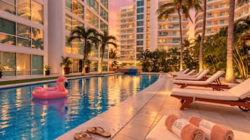 A swimming pool with lounge chairs and a flamingo float, surrounded by palm trees and modern buildings.