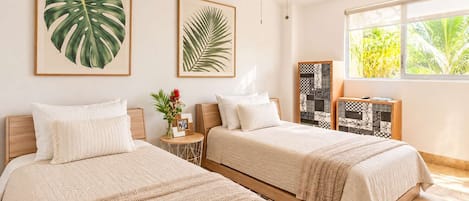 A bedroom with two beds, a ceiling fan, framed tropical leaves, a small table with a vase and picture, and a window with a view of greenery.