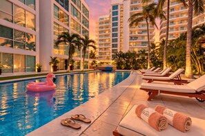 A swimming pool with lounge chairs and a flamingo float, surrounded by palm trees and modern buildings. - Coastal Comfort in Nuevo Vallarta (Bucerías)