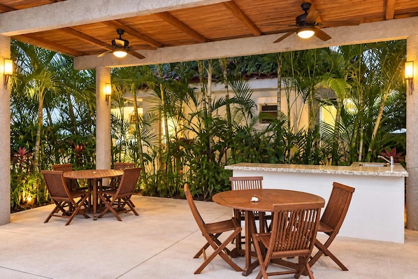 Un espace extérieur aménagé en terrasse, avec des tables et des chaises en bois, un ventilateur de plafond et un comptoir de bar.
